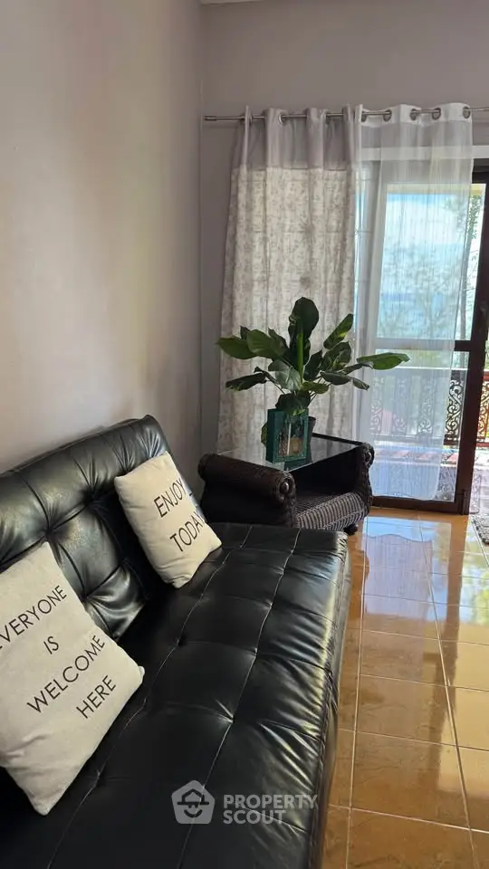 Cozy living room with black leather sofa, decorative pillows, and a view of the balcony through sheer curtains.