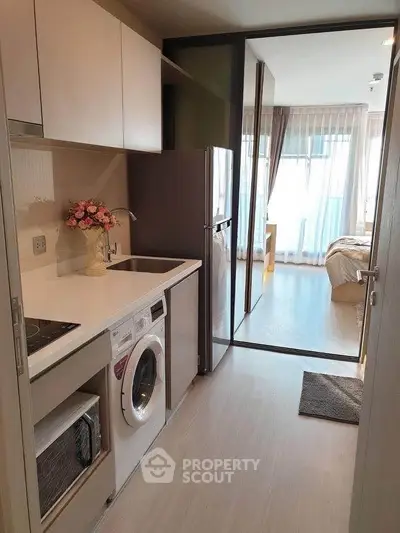 Modern compact kitchen with washing machine and open layout leading to a bright bedroom.