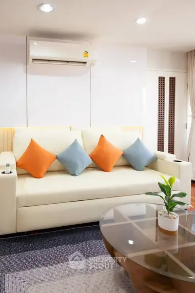 Modern living room with stylish sofa and vibrant cushions, featuring a sleek glass coffee table and air conditioning.
