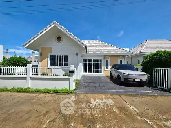 Charming single-story house with driveway and car, featuring a white picket fence and lush greenery.