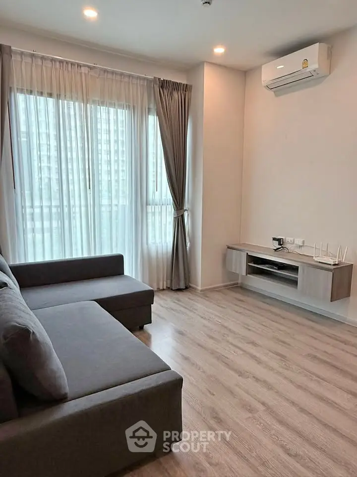 Modern living room with large window and air conditioning, featuring a sleek gray sofa and wooden flooring.