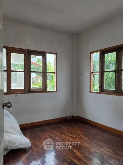 Bright empty room with wooden floors and large windows offering lush garden views.