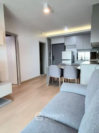 Modern living room with open kitchen and dining area, featuring sleek furniture and bright lighting.