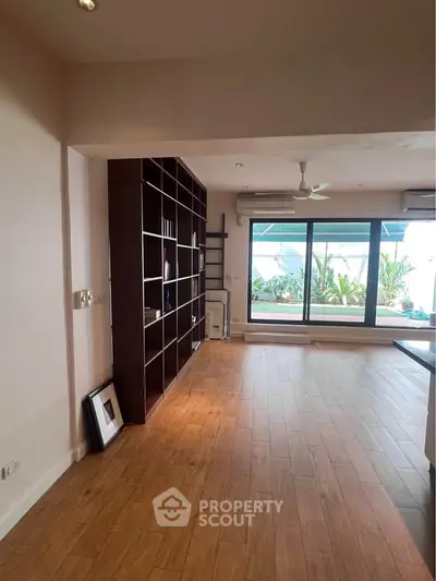 Spacious living room with wooden flooring and large windows opening to a balcony with greenery.