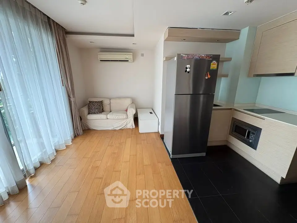 Modern living room with wooden flooring, cozy sofa, and sleek kitchen area with stainless steel fridge.