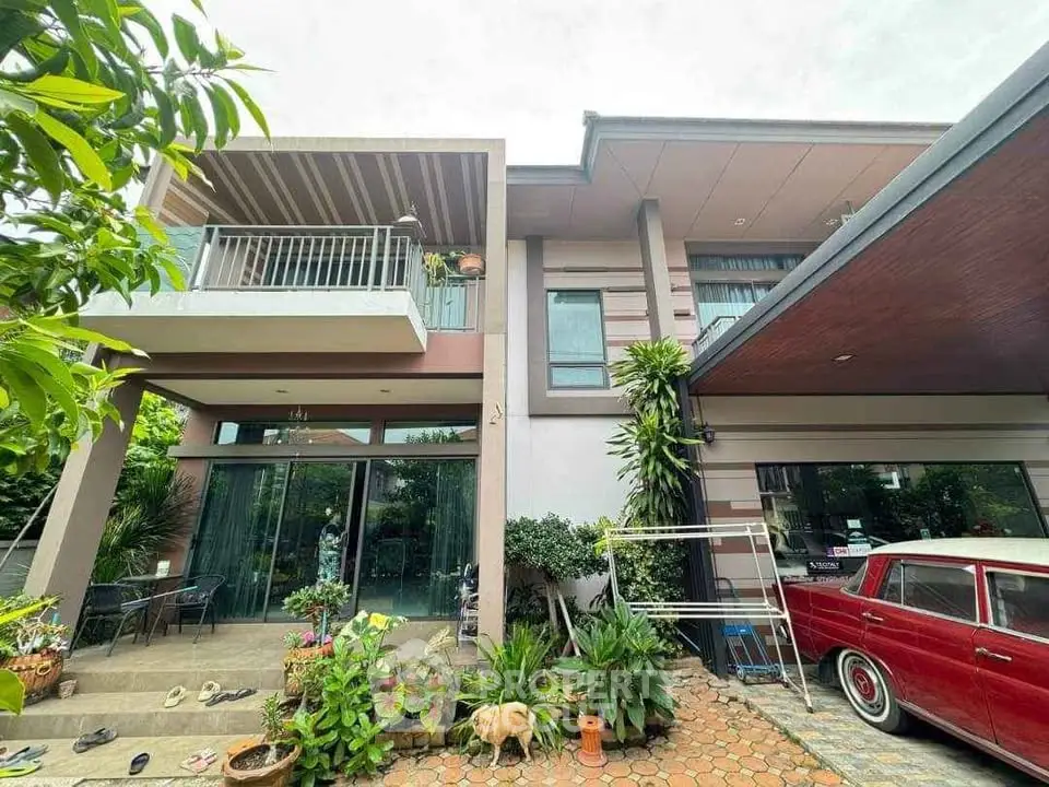 Modern two-story house with balcony and vintage car in driveway, surrounded by lush greenery.