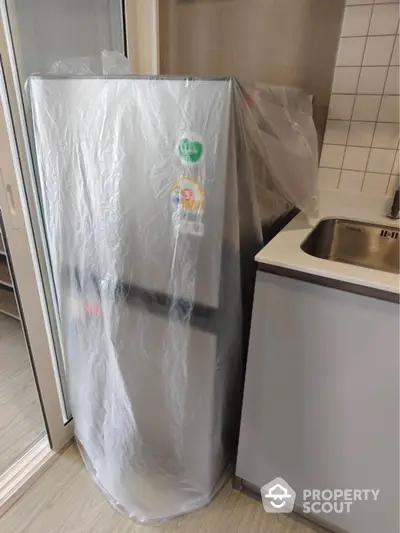 Modern kitchen with new fridge wrapped in protective plastic beside sleek sink.