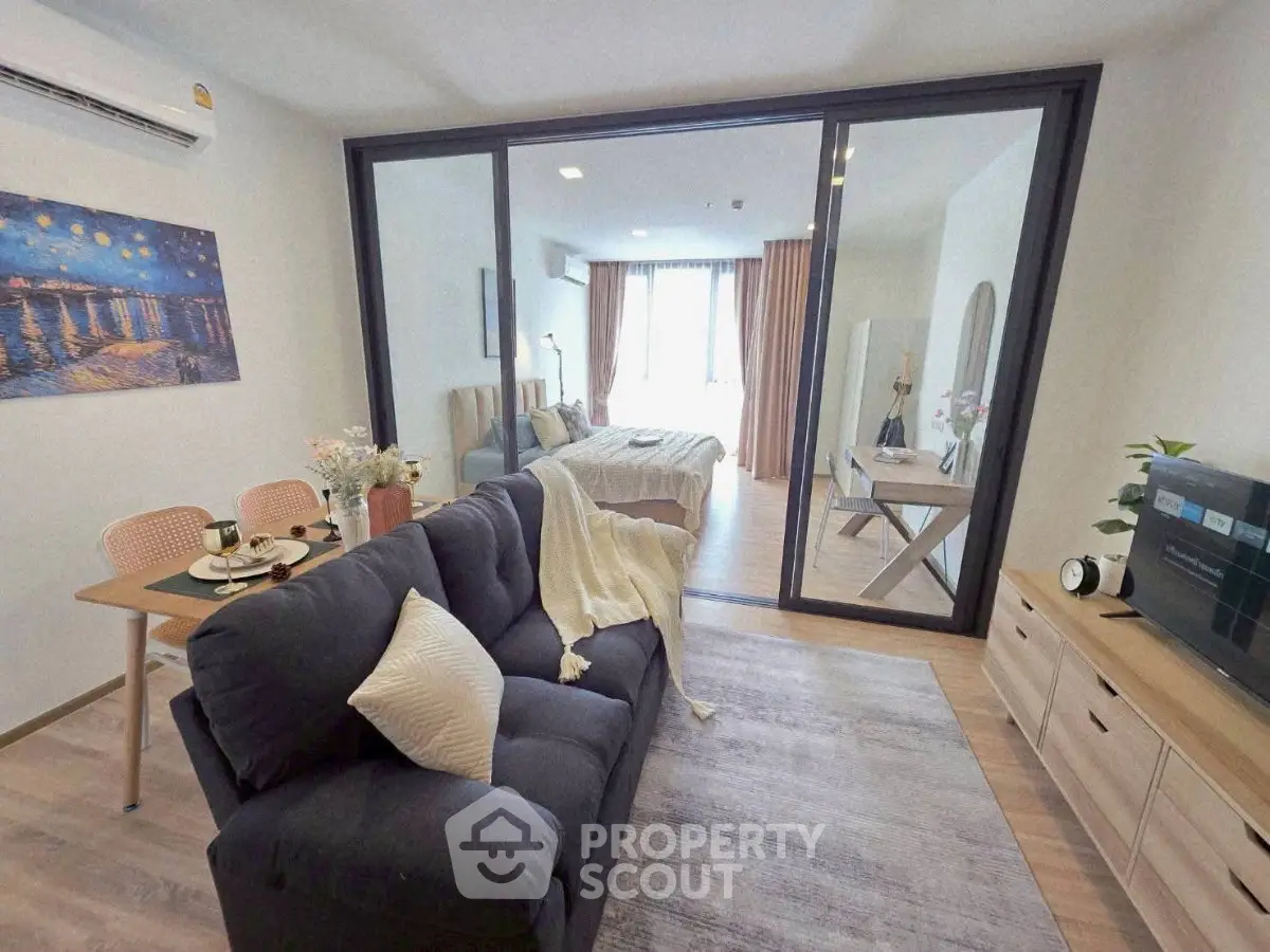 Modern open-plan living room with cozy seating and stylish decor, leading to a bright bedroom through glass doors.