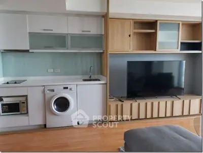 Modern apartment interior with compact kitchen and living area featuring a washing machine and large TV.