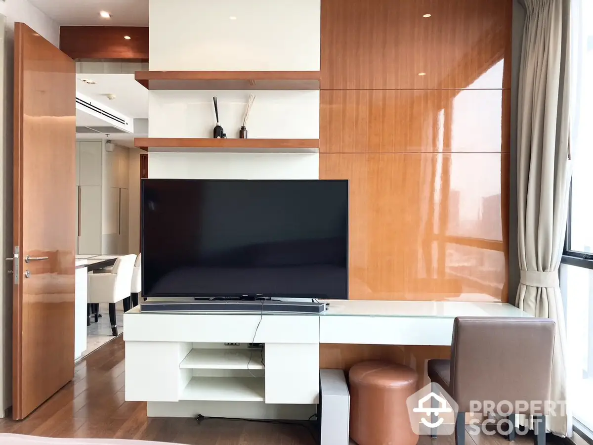 Modern living room with sleek furniture and wood paneling, featuring a large flat-screen TV and an open-plan layout connecting to the kitchen.