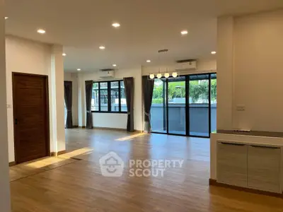 Spacious living room with hardwood floors, ample natural light, and modern lighting fixtures, leading to a cozy balcony with a serene view.