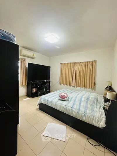 Cozy bedroom with large TV and air conditioning, featuring tiled flooring and neutral curtains.