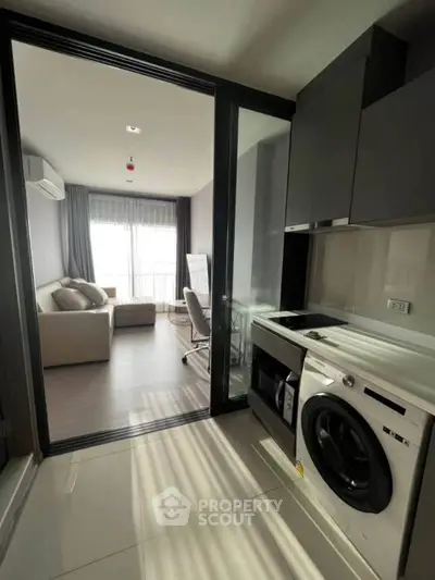 Modern apartment with sleek kitchen and cozy living room, featuring a washing machine and ample natural light.