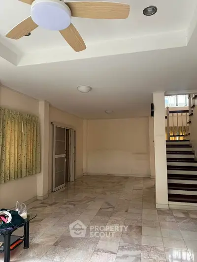 Spacious living room with marble flooring and ceiling fan, featuring a staircase and large window for natural light.