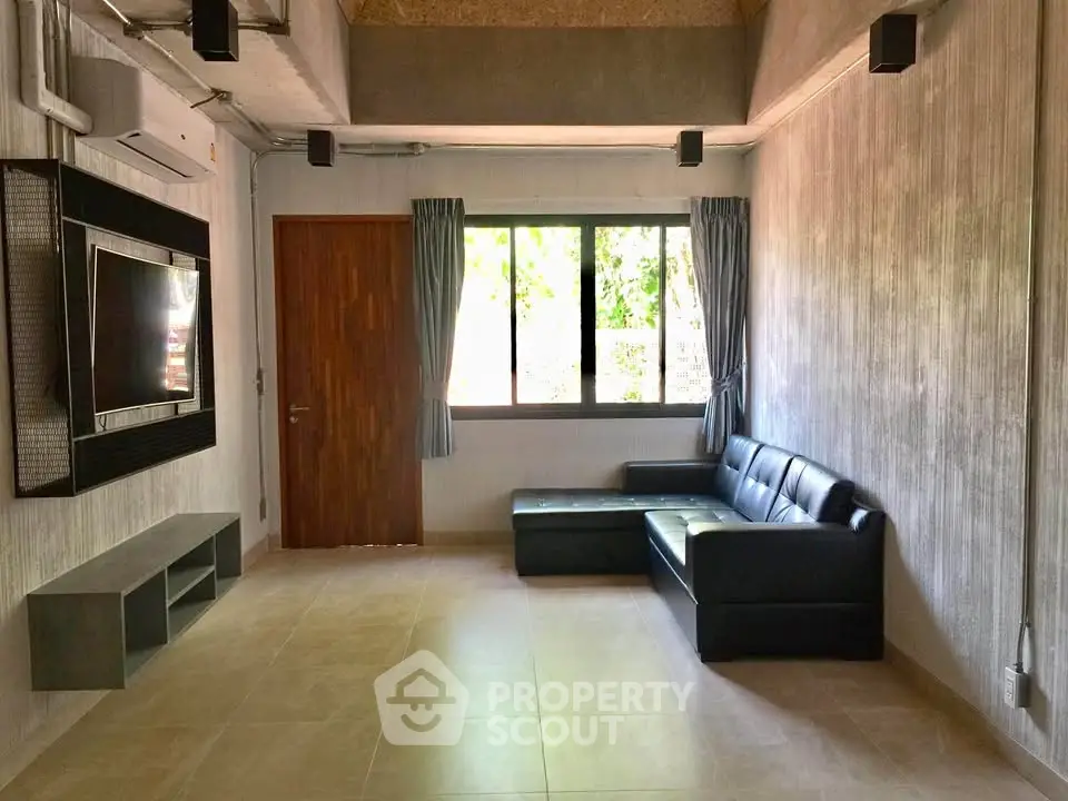 Modern living room with sleek black sofa and wall-mounted TV, featuring large window for natural light.