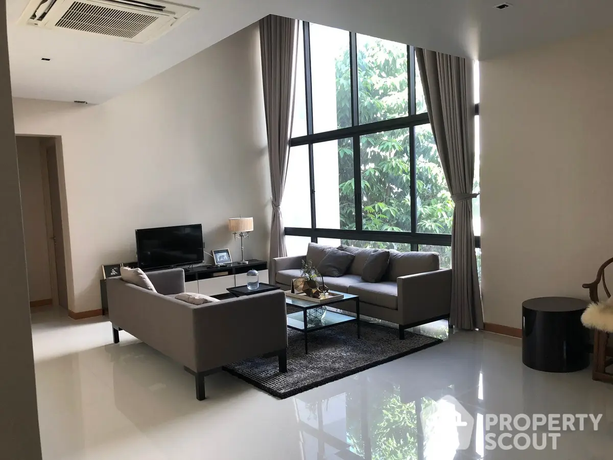 Spacious living room with high ceilings, large windows offering abundant natural light, and a serene view of greenery, complemented by modern furniture and a glossy tiled floor.