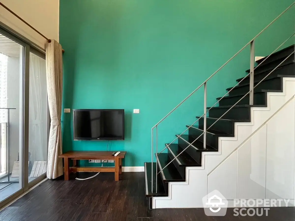 Spacious living area with striking green accent wall, modern staircase, and sleek hardwood floors, leading to a sunlit balcony.