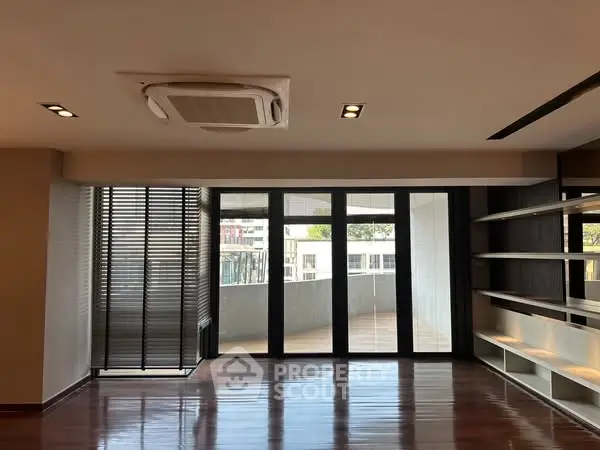 Spacious living room with large windows and wooden flooring, featuring built-in shelves and modern lighting.