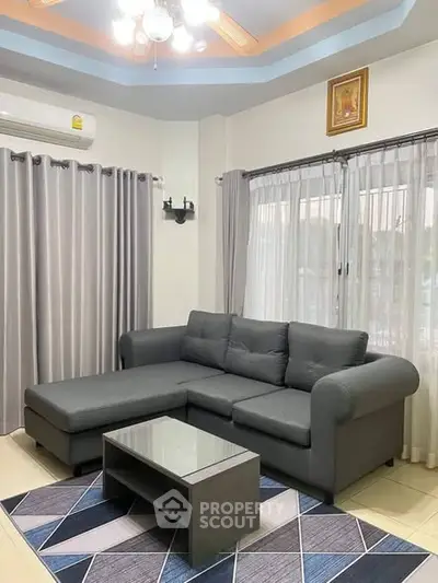 Modern living room with stylish gray sofa and glass coffee table, featuring elegant curtains and ceiling lighting.