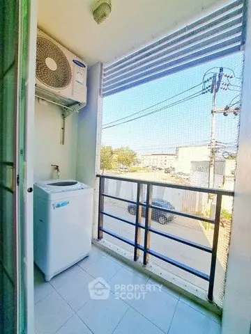 Compact balcony with washing machine and air conditioning unit, overlooking urban street view.