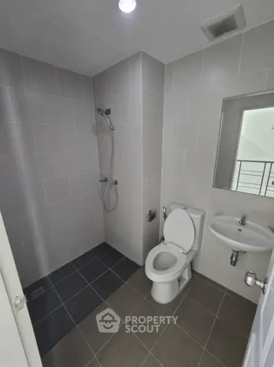 Modern bathroom with sleek gray tiles, wall-mounted shower, classic white toilet, and pedestal sink, featuring a small window for natural light.
