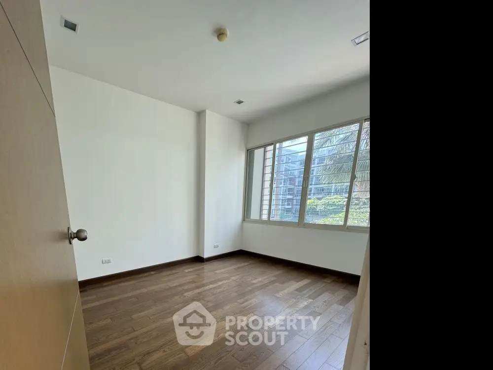 Spacious empty room with wooden flooring and large window offering natural light.