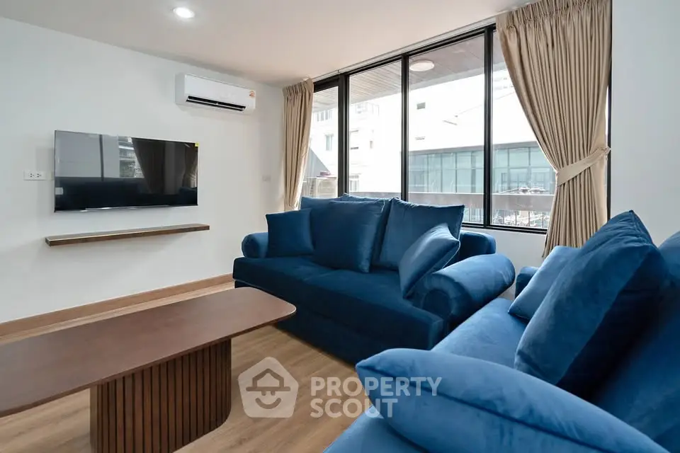 Modern living room with blue sofas, wall-mounted TV, and large windows for natural light.