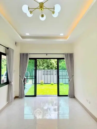 Bright living room with large windows overlooking a lush garden, featuring modern lighting and elegant curtains.