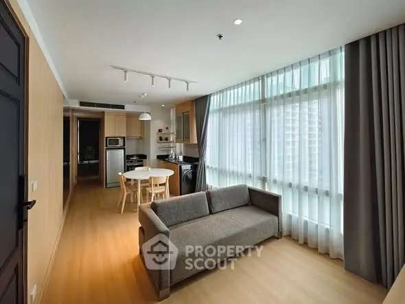 Modern open-layout living room with kitchen, featuring large windows and sleek furnishings.