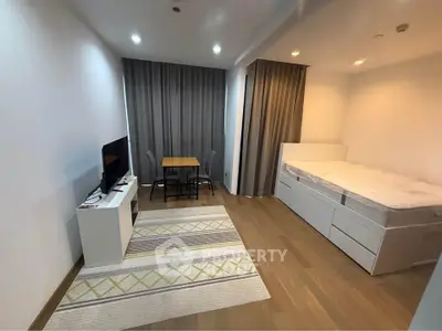 Modern studio apartment with cozy bed, TV, and dining area, featuring sleek wooden flooring and elegant curtains.