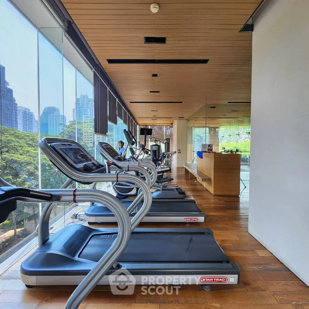 Modern gym with treadmills and city view, featuring floor-to-ceiling windows and wooden flooring.