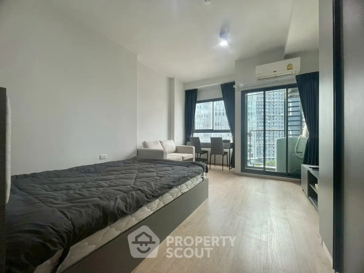 Modern studio apartment with city view, featuring cozy bed, seating area, and large windows for natural light.