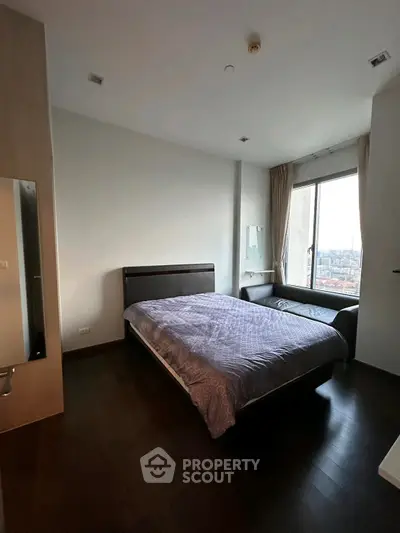 Modern bedroom with large window offering city view, featuring a cozy bed and sleek wooden flooring.