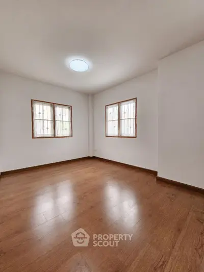 Spacious and well-lit empty room with polished hardwood flooring and dual windows offering ample natural light, ideal for a personalized living space.