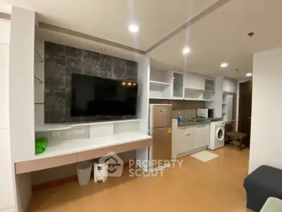 Modern open-layout living room with kitchen, featuring a large TV and stainless steel appliances.