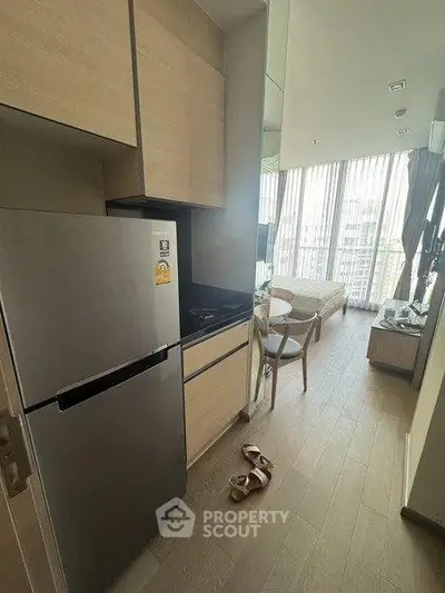 Modern compact kitchen with fridge and dining area in a stylish apartment with floor-to-ceiling windows.
