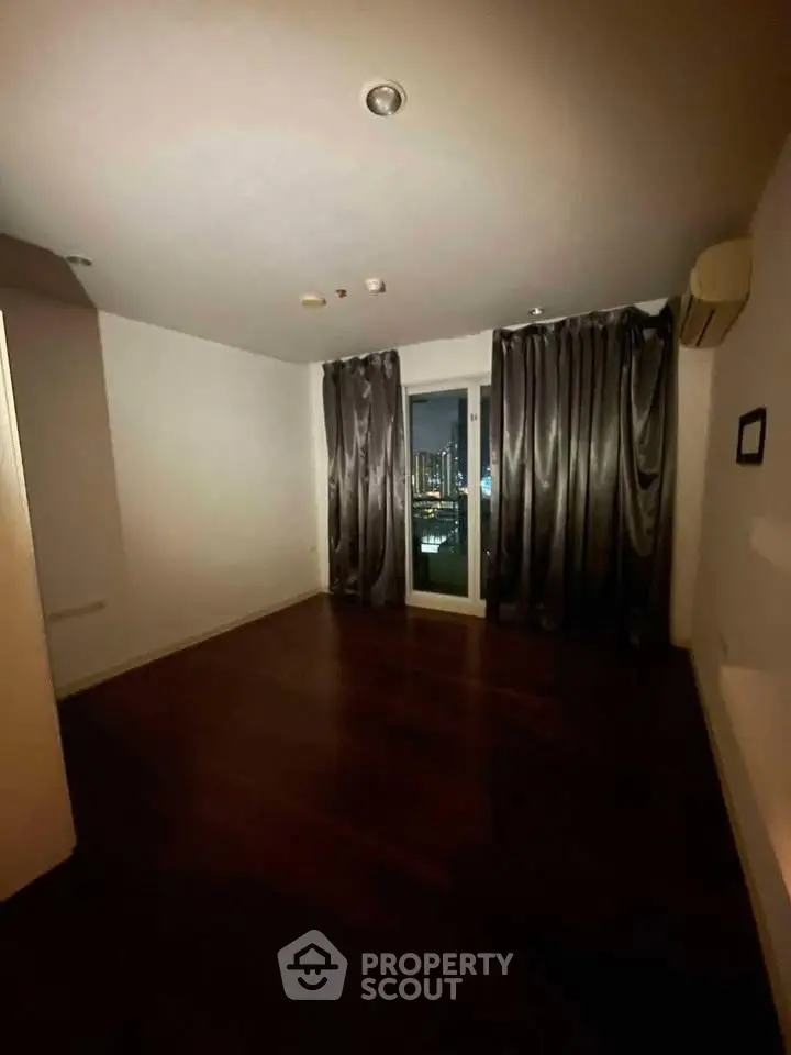 Spacious bedroom with dark wood flooring and city view from window.