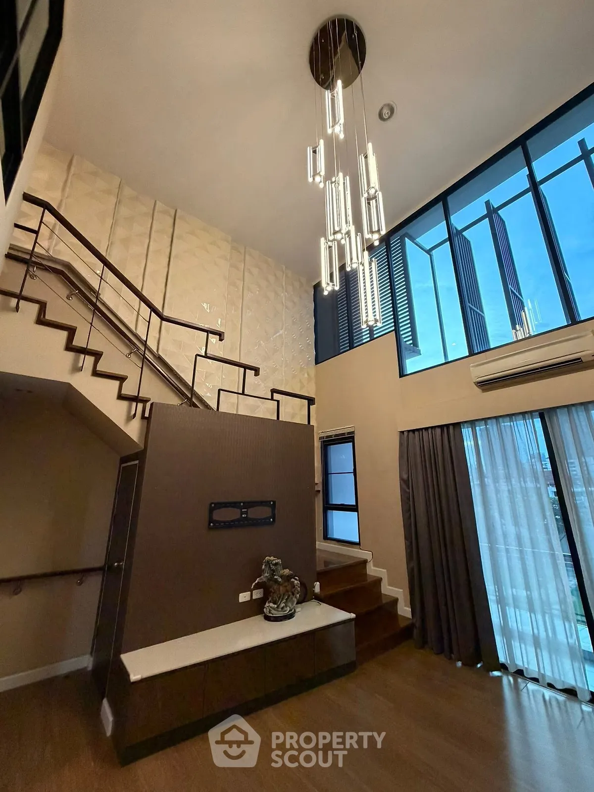 Luxurious modern living room with high ceilings and elegant staircase, featuring large windows and stylish lighting.