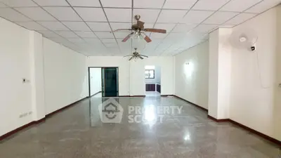 Spacious empty living room with ceiling fans and tiled floor, leading to a kitchen area.
