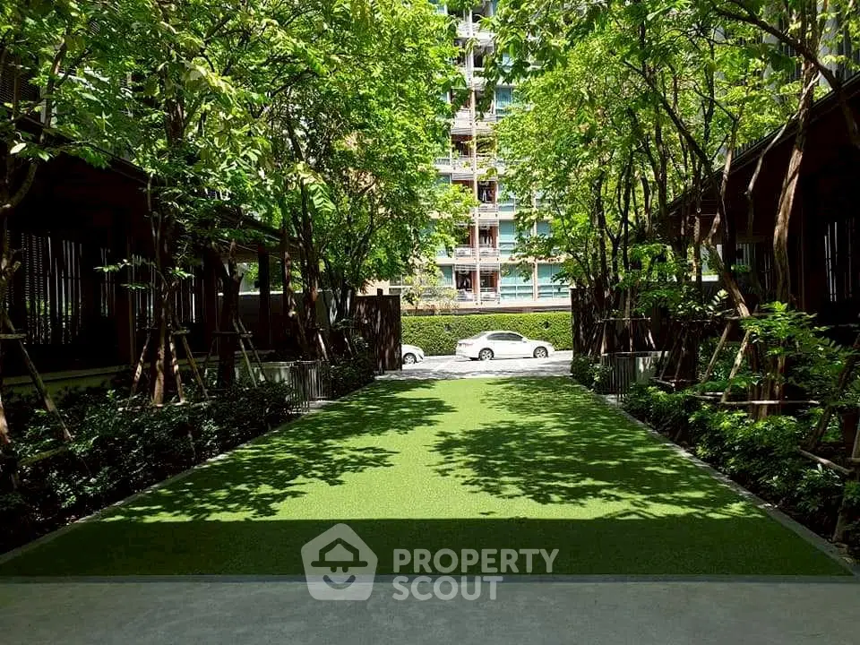 Lush green garden pathway in modern residential complex with trees and parked cars.
