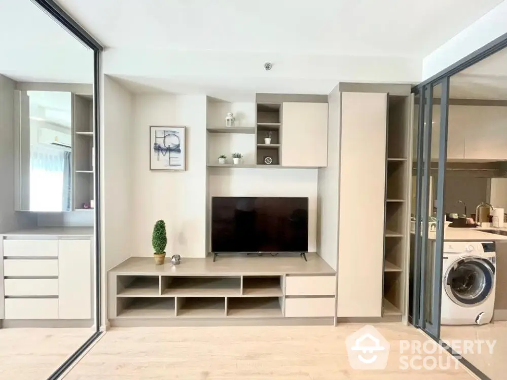 Modern apartment interior with integrated living space, featuring sleek built-in entertainment unit, adjacent to a well-equipped kitchen with washing machine.