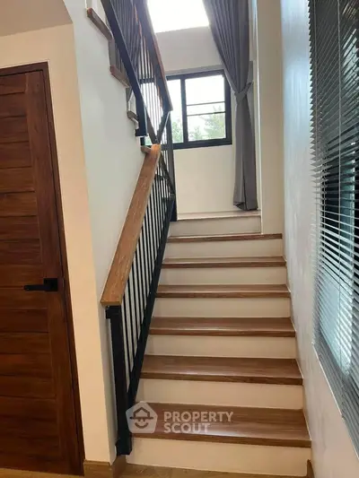 Elegant staircase with wooden steps and wrought-iron balusters, complemented by natural light from a window with stylish curtains.