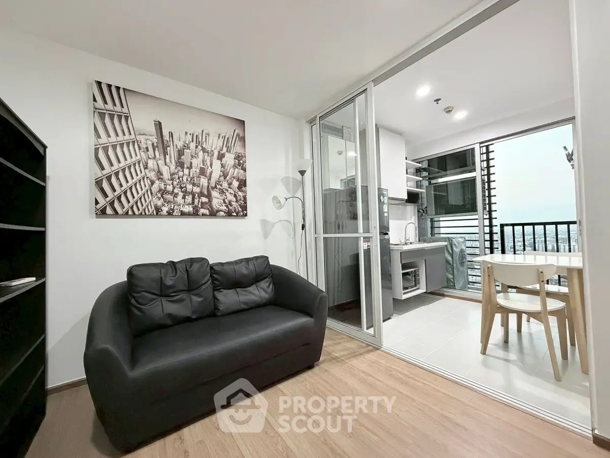 Modern living room with balcony and city view, featuring sleek black sofa and open kitchen layout.