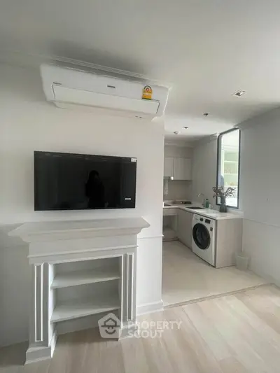 Modern living room with built-in TV and compact kitchen featuring washing machine.