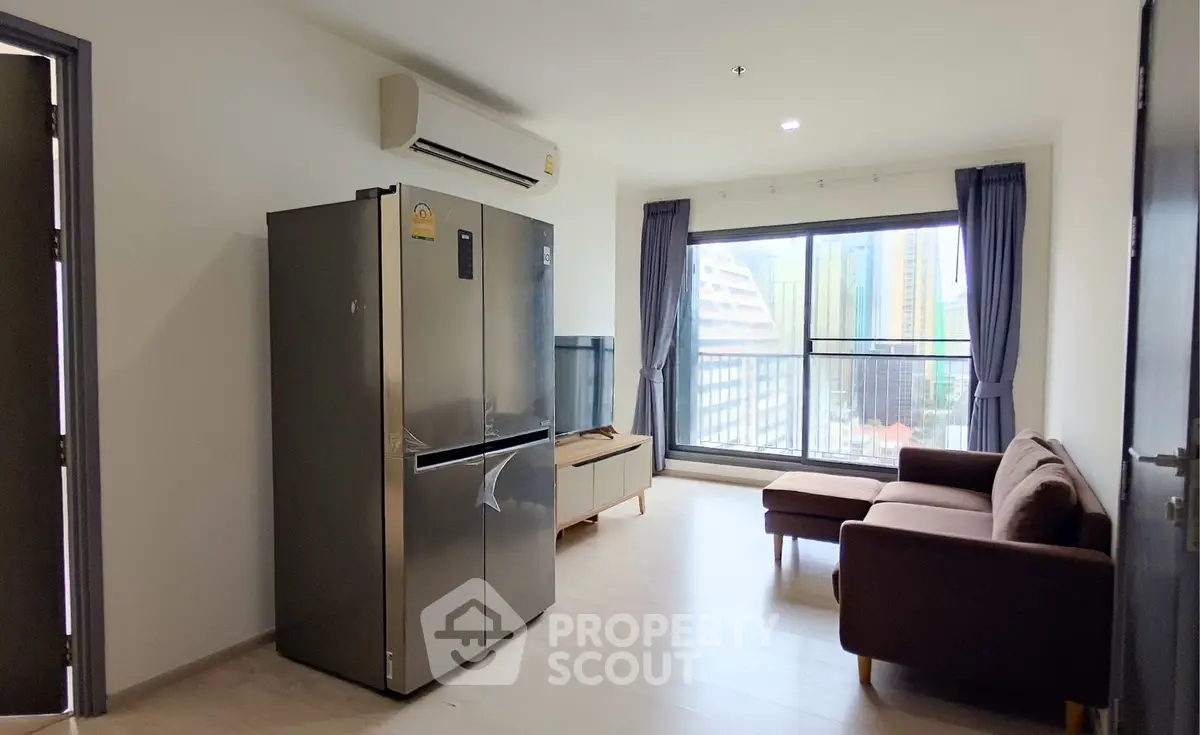 Modern living room with large window, brown sofa, and stainless steel refrigerator in a bright apartment.
