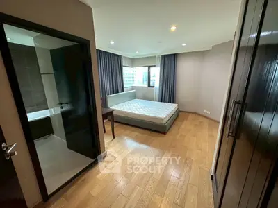 Spacious bedroom with wooden flooring and large window offering city views, adjacent to modern bathroom.