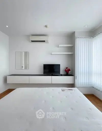 Modern minimalist bedroom with wall-mounted TV and air conditioning, featuring sleek white decor and natural light.