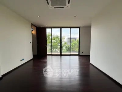 Spacious empty room with large windows and dark wood flooring, offering a serene view of greenery.