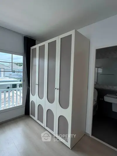 Modern bedroom with stylish wardrobe and partial bathroom view, featuring large window with natural light.