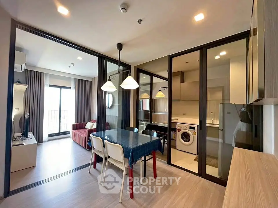 Modern open-plan living room with dining area and glass partitions, featuring a washing machine and stylish decor.